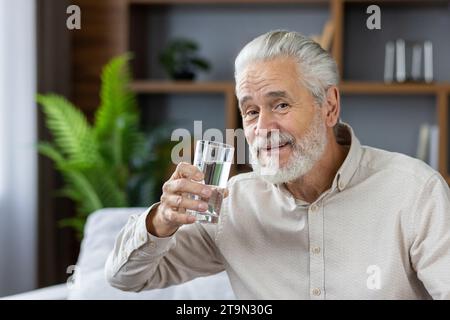 Portrait rapproché d'un homme âgé aux cheveux gris assis à la maison sur le canapé tenant un verre d'eau propre, menant un mode de vie sain, pointant vers la caméra et souriant. Banque D'Images