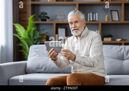 Homme âgé aux cheveux gris assis sur le canapé à la maison, utilisant une tablette, parlant lors d'un appel vidéo et regardant la caméra. Banque D'Images