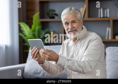 Portrait rapproché d'un homme âgé aux cheveux gris assis sur le canapé à la maison, tenant une tablette et souriant à la caméra. Banque D'Images