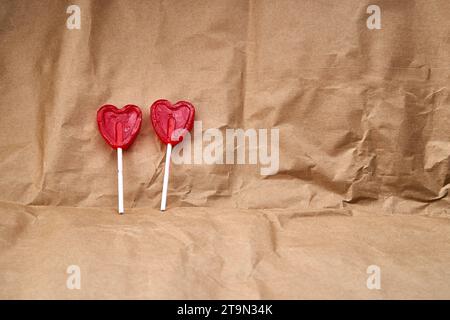 Deux sucettes rouges en forme de coeur représentant ensemble l'amour, sur un fond texturé en carton. Banque D'Images