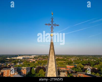 Capturée d'un point de vue aérien, cette image montre la flèche d'une église avec une croix ornée perchée au sommet, atteignant vers le vaste ciel bleu. La flèche est un symbole poignant de foi et de spiritualité, dominant le paysage urbain environnant. Le ciel clair en arrière-plan est strié de traînées délicates d'avions, faisant allusion à l'activité animée au-delà de ce sommet architectural paisible. Heavenward : Church Spire et Cross Against the Sky. Photo de haute qualité Banque D'Images