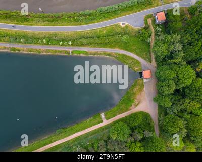 Capturée d'en haut, cette image montre un étang tranquille bordé par une route soigneusement pavée et des sentiers de randonnée qui se croisent. La verdure luxuriante empiétant sur les chemins crée un coin confortable au bord de l'eau, invitant un moment de pause. Les textures contrastées et l'harmonie entre les éléments artificiels et naturels évoquent un sentiment de coexistence pacifique dans ce coin tranquille du monde. Sérénité aérienne : sentiers et étang. Photo de haute qualité Banque D'Images