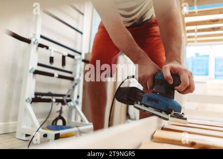 l'homme méconnaissable frotte le lit d'enfant en bois avec une ponceuse électrique ou un broyeur en enlevant la vieille peinture Banque D'Images