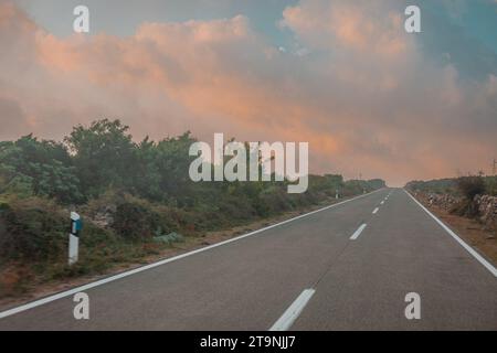 Route droite épique sur l'île de Cres avec des nuages colorés au-dessus d'elle, tôt le matin avec le lever du soleil. Peinture comme scène magique sur la route. Banque D'Images