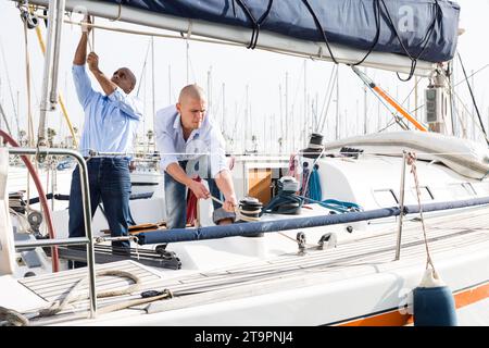 Deux hommes en chemises bleues et jeans travaillant sur un voilier dans le port Banque D'Images