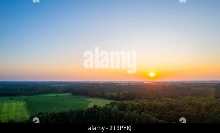 L'image capture la beauté sereine du lever du soleil sur un paysage rural tranquille. L'horizon est embrasé par les teintes douces et chaudes du soleil matinal, alors qu'il s'élève au-dessus d'une forêt dense et projette sa lueur sur les champs adjacents. Le ciel passe des oranges et jaunes ardents près du soleil aux bleus et violets frais de la nuit persistante, marquant le début d'une nouvelle journée. La vaste ouverture des champs contraste avec la canopée luxuriante de la forêt, créant une toile qui célèbre l'aube du possible. La nouvelle lumière de l'aube sur les champs. Photo de haute qualité Banque D'Images