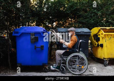 Homme senior en fauteuil roulant jetant des déchets de papier, du carton dans un conteneur de recyclage devant son appartement. Homme âgé triant les déchets selon Banque D'Images