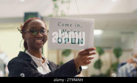 Employé afro-américain ouvrant le magasin de vêtements le matin, attendant les clients. Employé de vente au détail BIPOC tournant le panneau de message fermé sur la porte de la boutique de mode dans le centre commercial Banque D'Images