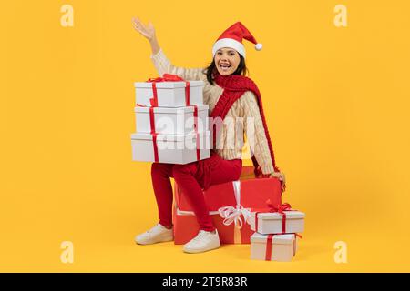 Femme assise sur pile de cadeaux de Noël emballés, fond jaune Banque D'Images