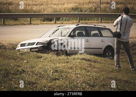 L'homme tire sur la voiture cassée sur une route de banlieue avec une caméra vidéo Banque D'Images