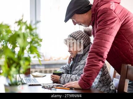 Couple senior en vêtements d'hiver passant sur leurs factures d'énergie sur ordinateur portable à la maison Banque D'Images