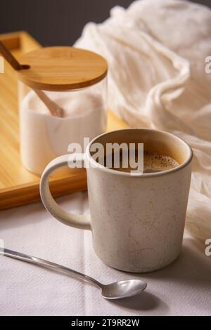 Tasse de café noir servi sur une tasse blanche. Il y a une soucoupe avec du sucre blanc sur un plateau en bois, et tout repose sur une nappe blanche. Banque D'Images