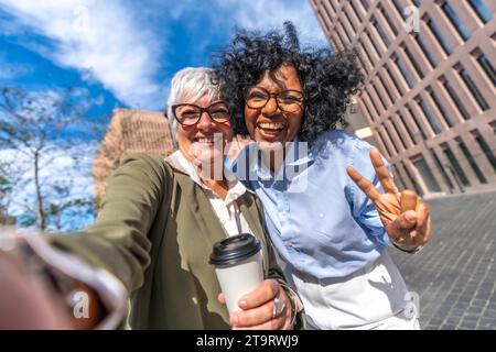 Photo frontale de deux femmes d'affaires souriantes multi-ethniques prenant selfie et faisant signe de victoire avec les doigts à l'extérieur Banque D'Images