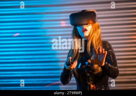 Femme blonde jouant à l'aide de lunettes de réalité Virtutal dans un espace de nuit urbain avec des lumières de néon Banque D'Images