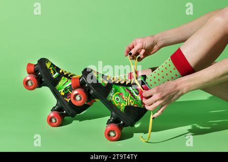 Vue de côté de la récolte anonyme femelle nouant des lacets et mis sur des lames de rouleau rétro colorées contre le mur vert tout en essayant le patinage à roulettes Banque D'Images