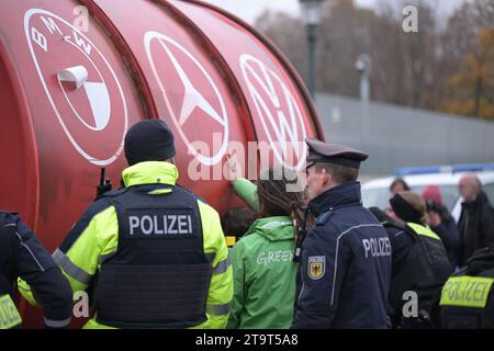 Berlin, Allemagne. 27 novembre 2023. Les policiers se tiennent devant un grand tonneau portant les logos des constructeurs automobiles BMW (l-r), Mercedes-Benz et Volkswagen. Le baril a été conduit devant la Chancellerie fédérale par des militants de l’organisation environnementale Greenpeace avant le sommet de la RCA. La chancelière Scholz rencontre des représentants de l’industrie automobile pour discuter de l’expansion de l’électromobilité sur les routes allemandes. Crédit : Hannes P Albert/dpa/Alamy Live News Banque D'Images