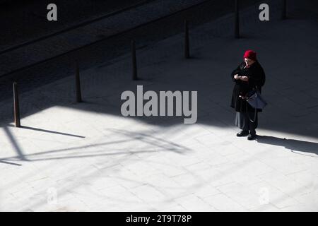 Belgrade, Serbie- 18 mars 2022 : une femme âgée avec un chapeau rouge debout seule sur une rue de la place de la ville par la journée froide et ensoleillée, vue à grand angle Banque D'Images