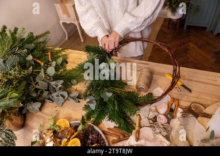 Vue de dessus composition de mains femelles faisant couronne de Noël à partir de branches naturelles d'épinette, de pin, d'eucalyptus. Fille enroule des brindilles vertes sur la base Kraft Banque D'Images