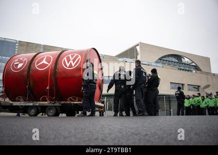 Berlin, Allemagne. 27 novembre 2023. Les policiers se tiennent devant un grand tonneau portant les logos des constructeurs automobiles BMW (l-r), Mercedes-Benz et Volkswagen. Le baril a été conduit devant la Chancellerie fédérale par des militants de l’organisation environnementale Greenpeace avant le sommet de la RCA. La chancelière Scholz rencontre des représentants de l’industrie automobile pour discuter de l’expansion de l’électromobilité sur les routes allemandes. Crédit : Hannes P Albert/dpa/Alamy Live News Banque D'Images