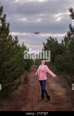 Jeune femme en rose tricoté à la campagne parmi les conifères dans la forêt posant pour une caméra drone dans le ciel. Frisé fille s'amusant dans nat Banque D'Images