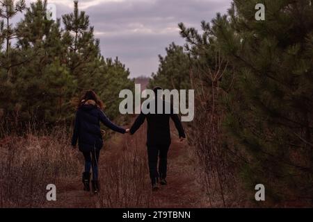 Heureux couple courant à travers la forêt se tenant la main. Un homme en manteau noir et une jeune femme en pull tricoté se baladent dans un parc de pins, pla Banque D'Images