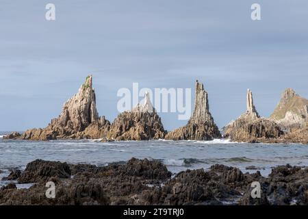 côte rocheuse avec trois formations rocheuses éminentes et dentelées dans l'océan, sous un ciel clair. Le premier plan montre des roches sombres, couvertes de barnacle Banque D'Images