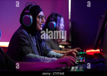 Portrait vue de côté de jeune femme musulmane jouant à des jeux vidéo dans le club de cybersports et souriant joyeusement, espace de copie Banque D'Images