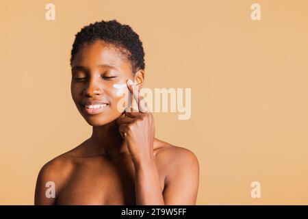 Jeune femme souriante noire les yeux fermés appliquant une crème hydratante sur son visage. Jeune femme traitant sa peau du visage avec un produit de beauté. Banque D'Images