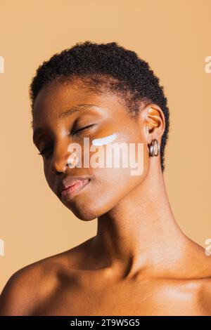 Jeune femme noire les yeux fermés avec une crème hydratante sur le visage. Jeune femme traitant sa peau du visage avec un produit de beauté. Banque D'Images