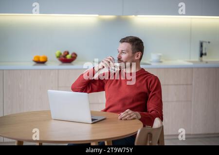 Homme freelance boire de l'eau ayant une pause dans le travail assis à table sur la cuisine à la maison avec ordinateur portable. Banque D'Images