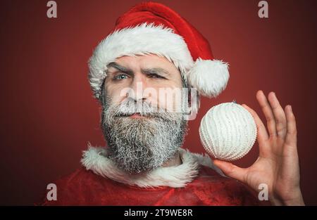 Père Noël avec barbe gelée avec boule de Noël blanche. Homme barbu dans les vêtements du Père Noël avec boule de jouet décoratif du nouvel an. Décorations de vacances et Banque D'Images