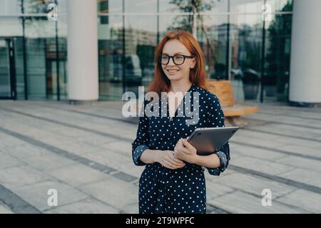 Femme d'affaires aux cheveux roux avec des lunettes tenant une tablette à l'extérieur d'un immeuble de bureaux moderne, souriant Banque D'Images