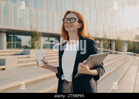 Femme d'affaires optimiste avec café et tablette marchant par un bureau, prêt pour la journée à venir Banque D'Images