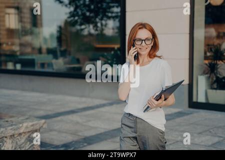 Femme aux cheveux roux dans des lunettes parlant au téléphone, tenant un dossier, à l'extérieur d'un bâtiment moderne Banque D'Images