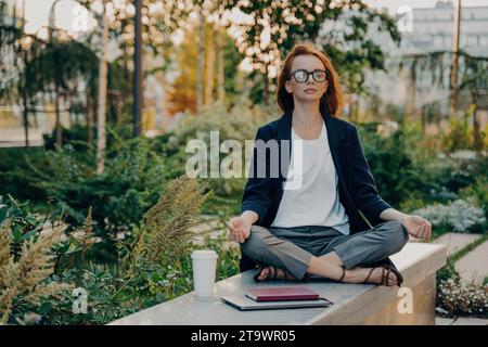 Femme d'affaires sereine méditant dans un parc avec un café et un cahier, trouver l'équilibre Banque D'Images