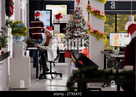 Équipe d'affaires travaillant dans un bureau décoré festif occupé pendant la saison des fêtes de Noël. Employés qui travaillent dur pour résoudre diverses tâches de projet d'entreprise dans l'espace de travail orné de Noël Banque D'Images