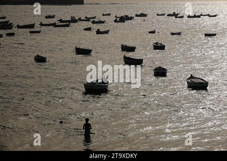 Un jeune garçon est silhouetté par le soleil couchant alors qu'il pataugeait dans l'eau au petit port de bateau de pêche dans la baie de Cadix le long du front de mer à Cadix, en Espagne. Banque D'Images