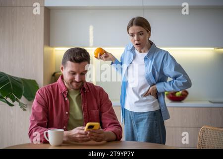 Émerveillé jaloux femme choquée se tient derrière le dos peeping dans l'homme mari smartphone sur la cuisine à la maison Banque D'Images