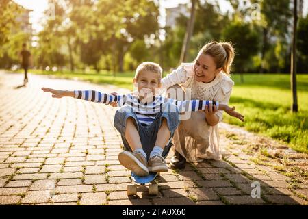 Mère s'amusant avec son fils dans le parc le jour ensoleillé. Garçon est en train de rider le skateboard et sa mère le pousse. Banque D'Images