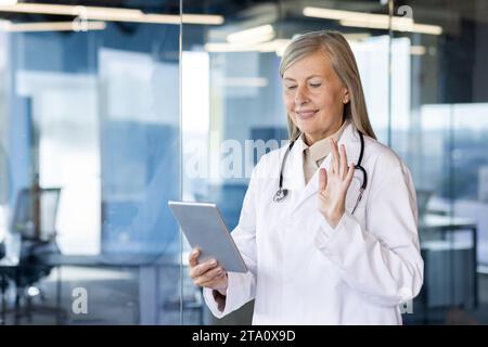 Une femme plus âgée aux cheveux gris se tient debout dans le bureau de la clinique dans un manteau, tenant un comprimé dans ses mains. clavardage lors d'un appel vidéo, mène une consultation en ligne, salue et fait des vagues à la caméra. Banque D'Images