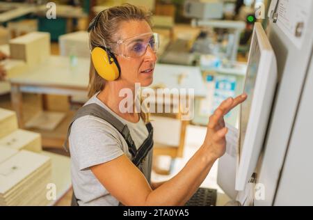 Femme avec protection auditive utilisant un écran tactile sur une machine de découpe à la menuiserie Banque D'Images