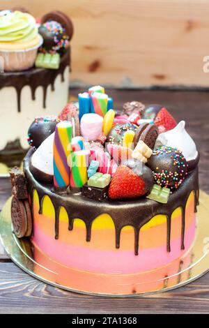 Gâteau rose dégradé richement décoré coloré avec des bonbons, des popsicles, des sucettes, de la gelée, des fraises et des cupcakes sur fond rustique Banque D'Images