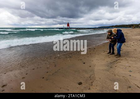 Marcheurs à la recherche de pierres de petoskey sur la plage de Charlevoix. Phare de South Pier en arrière-plan. Journée orageuse sur la plage de Charlevoix avec le phare de Charlevoix South Pier en arrière-plan. On trouve des pierres de Charlevoix et de Petoskey sur la plage de Charlevoix, surtout par vent fort et houle. Charlevoix, États-Unis Banque D'Images