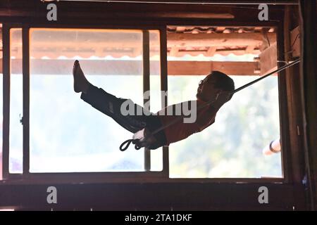 Rongshui. 28 novembre 2023. Un garçon joue sur le swing dans un bâtiment sur pilotis dans le village de Wuying à la frontière entre la région autonome de Guangxi Zhuang dans le sud de la Chine et la province du Guizhou dans le sud-ouest de la Chine, 4 octobre 2023. Wuying Village est un hameau du groupe ethnique Miao qui se niche confortablement dans les montagnes imposantes qui s'étendent à travers la frontière entre Guangxi et Guizhou. La plupart des villageois vivent encore dans des bâtiments traditionnels sur pilotis en bois. Crédit : Huang Xiaobang/Xinhua/Alamy Live News Banque D'Images