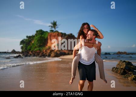 Couple heureux aime lune de miel sur la plage tropicale. L'homme porte la femme piggyback avec joie. Pieds nus sourires, marche le long du rivage. Amour, romance fleurir Banque D'Images