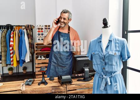 Homme d'âge moyen à cheveux gris assistant de magasin utilisant l'ordinateur parlant sur le smartphone à la boutique de vêtements Banque D'Images