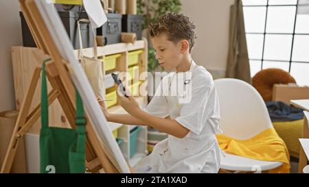 Adorable artiste garçon blond regardant sérieusement le smartphone, apprenant à dessiner dans la classe de studio d'art confortable Banque D'Images