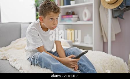 Adorable enfant blond portant une expression ennuyée, se bousculant confortablement sur le canapé à la maison, semblant sérieux tout en se relaxant et en regardant la télévision dans le salon Banque D'Images