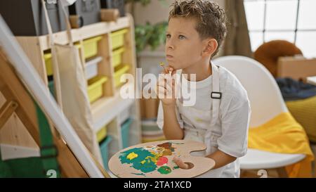 Adorable artiste garçon blond, dessinant avec confiance dans un studio d'art intérieur, regardant la toile avec soin, vêtu d'un tablier et d'un pinceau à la main. Banque D'Images