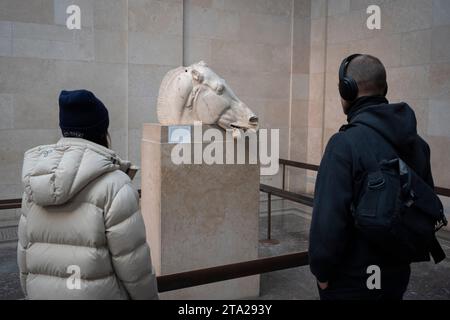 Les visiteurs du British Museum de Londres admirent la tête du cheval, l'une des métopes grecques antiques du Parthénon aussi connue sous le nom de marbres du Parthénon (Elgin), le 28 novembre 2023, à Londres, en Angleterre. La tête du cheval tirait autrefois le char de la déesse de la lune Selene. Quatre-vingt-douze métopes étaient des dalles de marbre rectangulaires placées sur les colonnes du temple du Parthénon d'Athènes représentant des scènes de la mythologie grecque. Banque D'Images